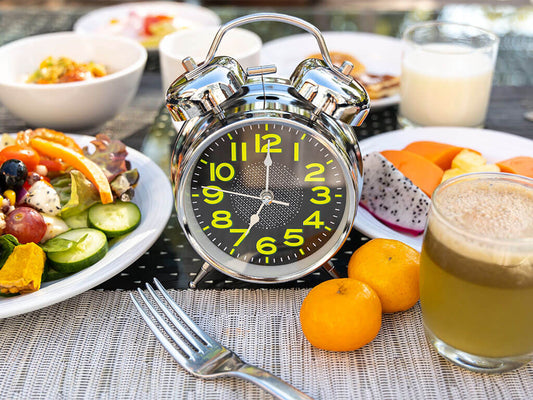 A silver analog alarm clock stands on a table among various breakfast items, including salad, fruit, juice, and a glass of milk.