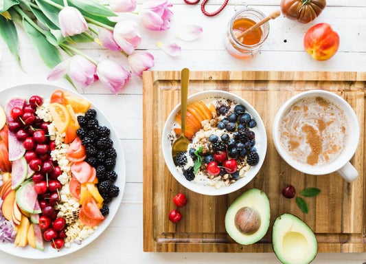 A breakfast scene with a fruit and cereal bowl, a plate with various fresh fruits, an avocado, a cup of coffee, honey, tulips and a peach on a wooden surface.