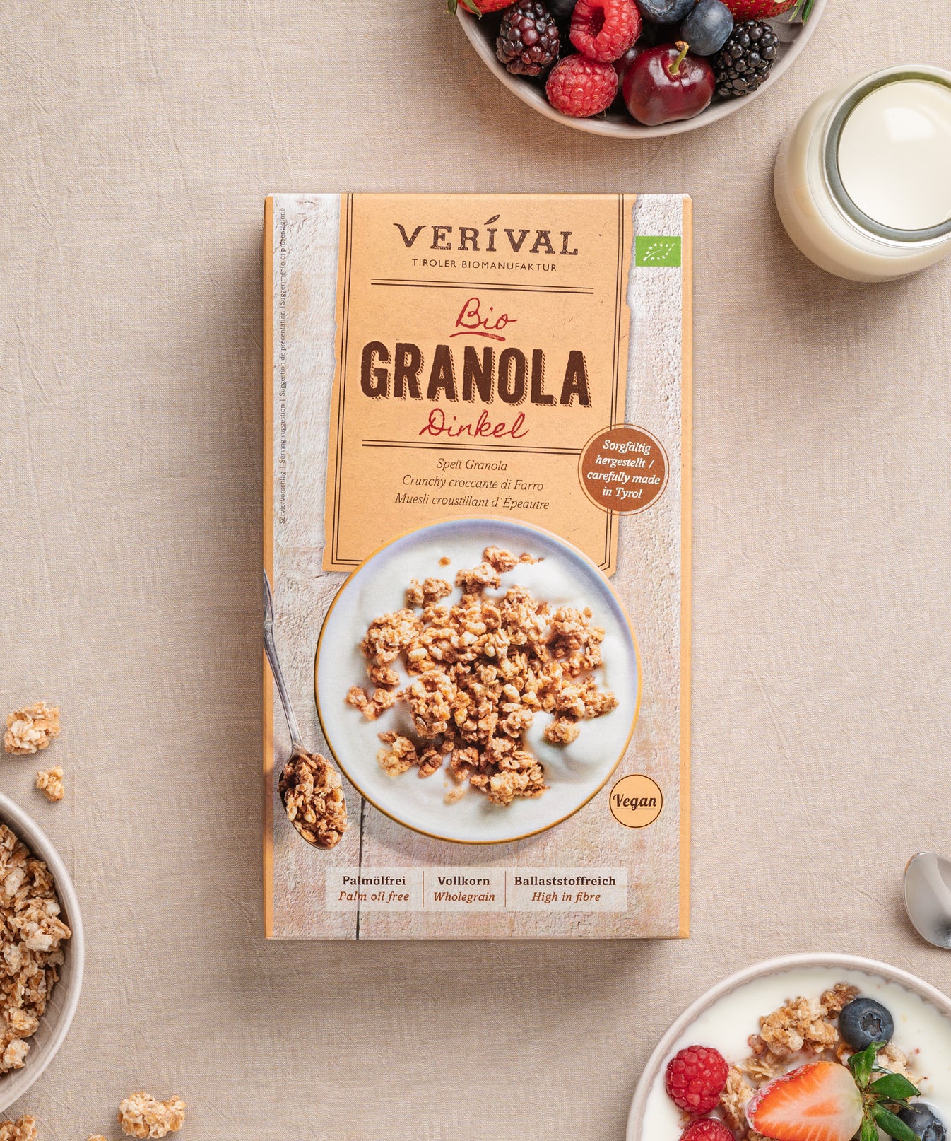Eine Packung Dinkel-Granola von Verival Bio steht mit Granola, Beeren, Milch und einem Löffel auf dem Tisch. Reich an Ballaststoffen eignet es sich perfekt für ein Knusper-Frühstück und ist die ideale vegane Wahl für gesundheitsbewusste Morgen.