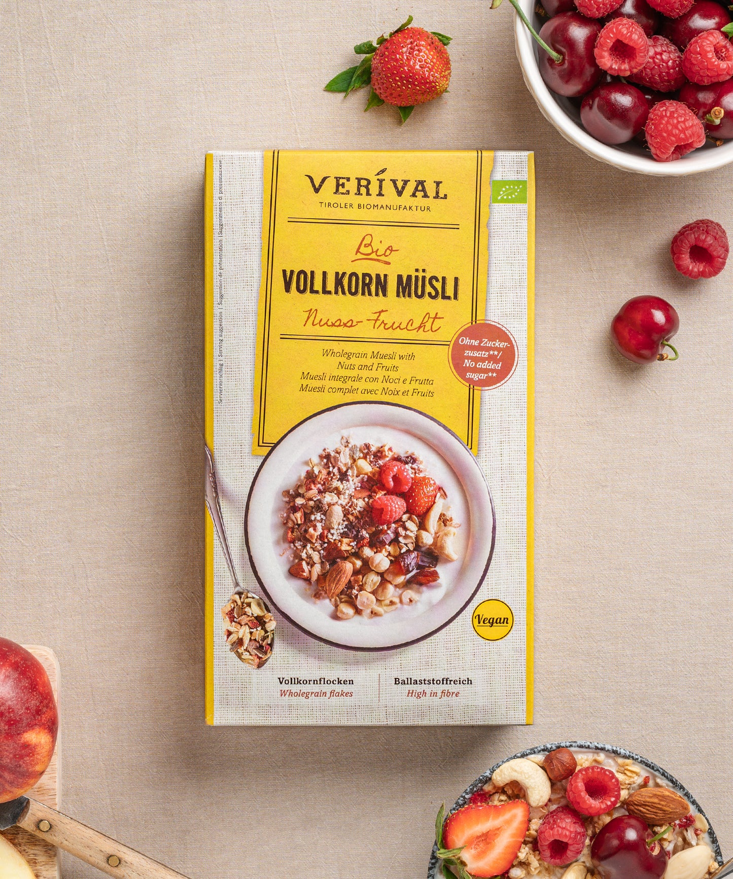 Eine Packung Nuss-Frucht Vollkorn Müsli von Verival Bio steht auf einem Tisch mit verschiedenen Früchten, geschnittenen Pfirsichen und Kirschen. Diese vegane Frühstücksoption ist köstlich und ballaststoffreich – perfekt für alle, die Wert auf eine nahrhafte Alternative legen.