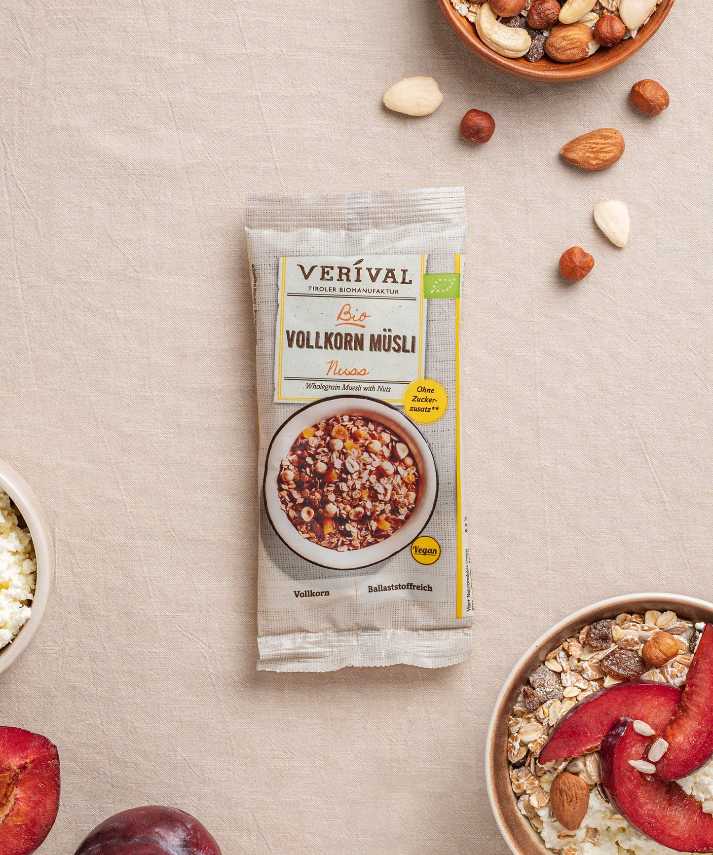 Eine Packung Nuss Vollkorn Müsli der Verival Bio GmbH liegt auf einer beigen Oberfläche, umgeben von verstreuten Nüssen, einer Schale mit Obst und Müsli sowie einem Apfel, und zeigt die nahrhaften Vorteile eines ballaststoffreichen veganen Frühstücks.