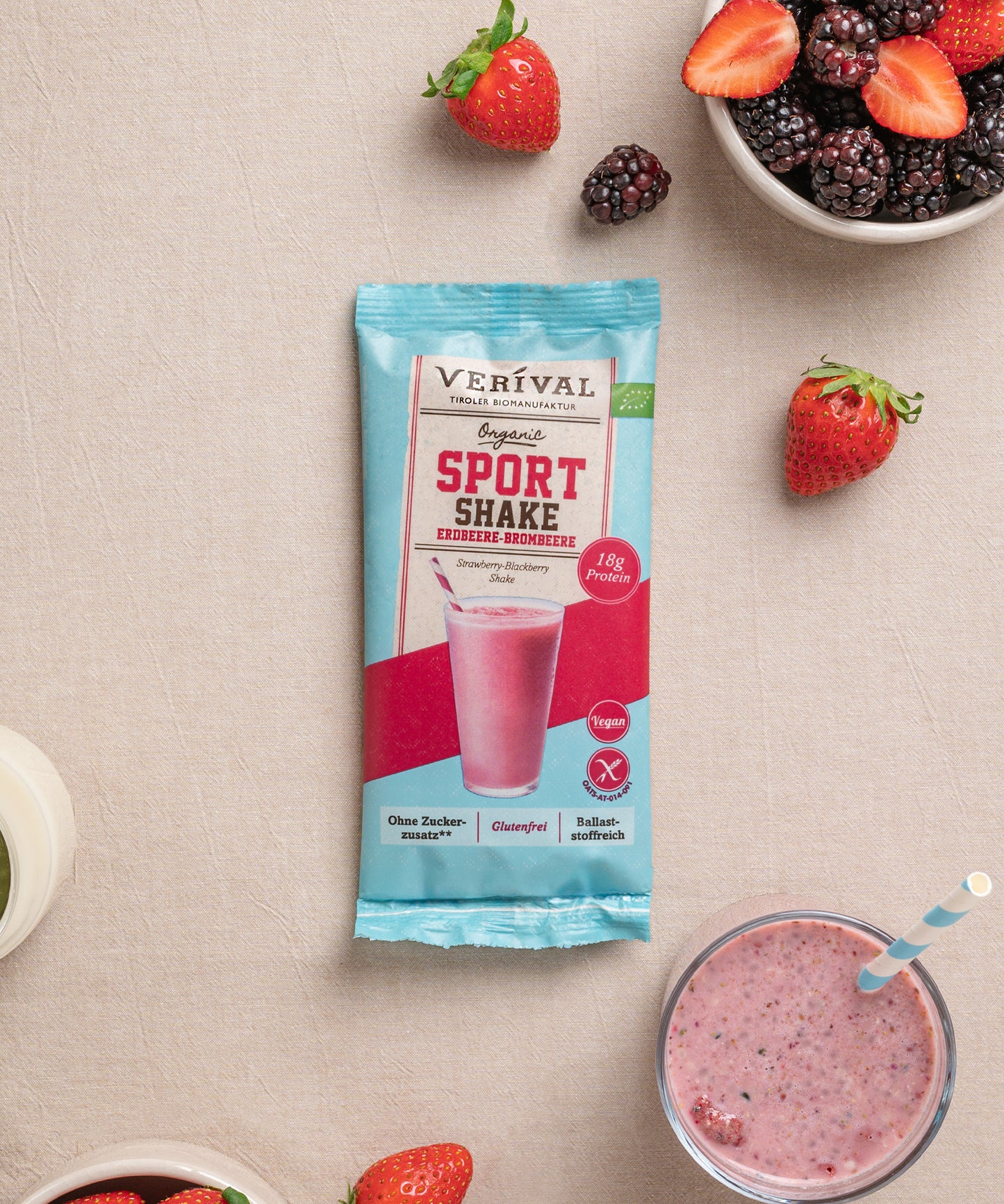 Eine Einzelportionspackung vom Sport Shake Erdbeere-Brombeere von Verival Bio ist umgeben von Erdbeeren, Brombeeren und dem Shake in einem Glas auf einem beigen Untergrund.