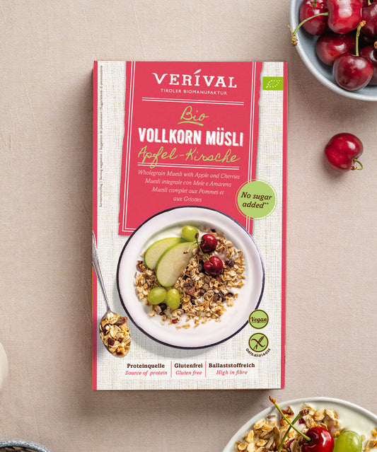 Eine Packung Apfel-Kirsch Vollkorn Müsli von Verival Bio, neben frischen Kirschen und einem Löffel Müsli. Die Packung informiert über den hohen Ballaststoffgehalt und die Freiheit von zusetztem Zucker.