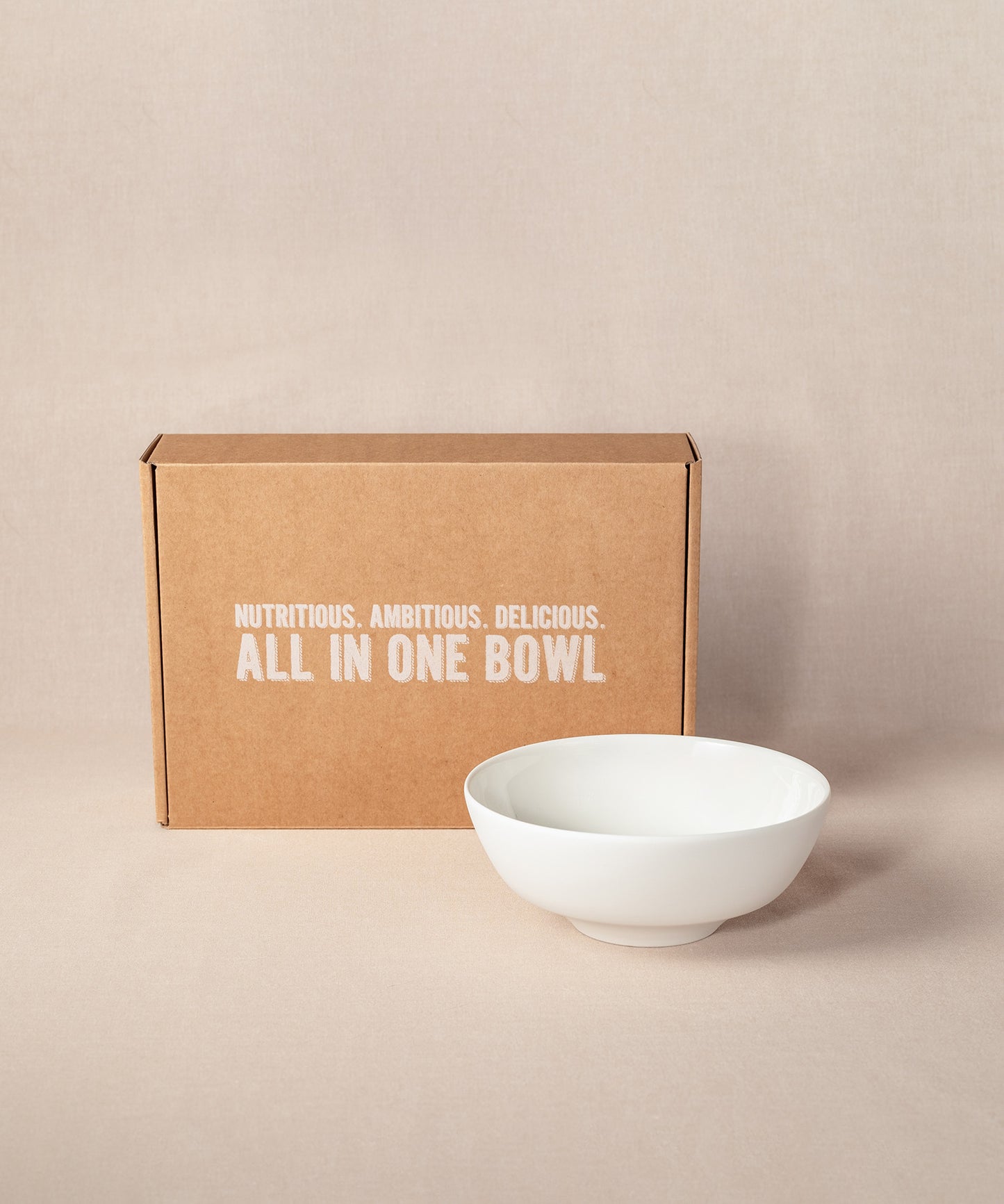 Eine braune Kartonschachtel mit dem Text „Nutritious. Ambitious. Delicious. All in one bowl“ steht aufrecht, neben einer eleganten, weißen Porridge-Schüssel aus Keramik. Die Schüssel ist Bestandteil des Sets „Breakfast Lover“ von Verival Bio.