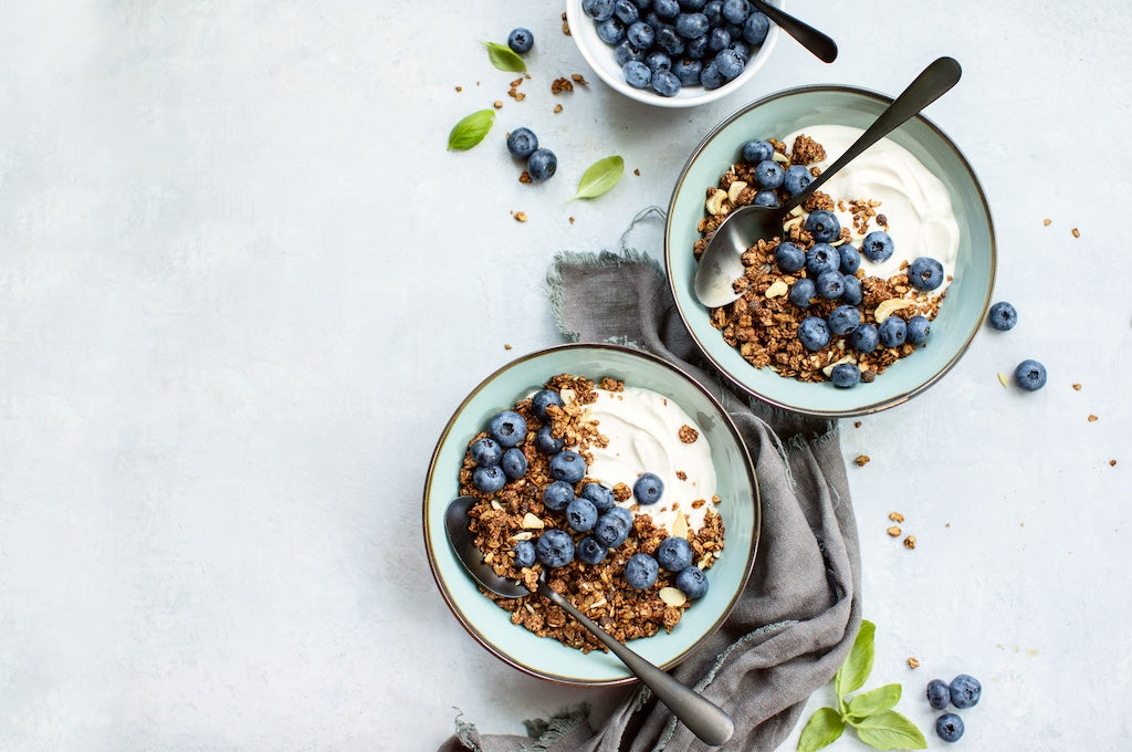 Zwei Schüsseln Joghurt mit Müsli und frischen Blaubeeren, in jeder Schüssel ein Löffel. Auf dem Tisch sind außerdem eine kleine Schüssel mit Heidelbeeren und verstreuten Blättern zu sehen.