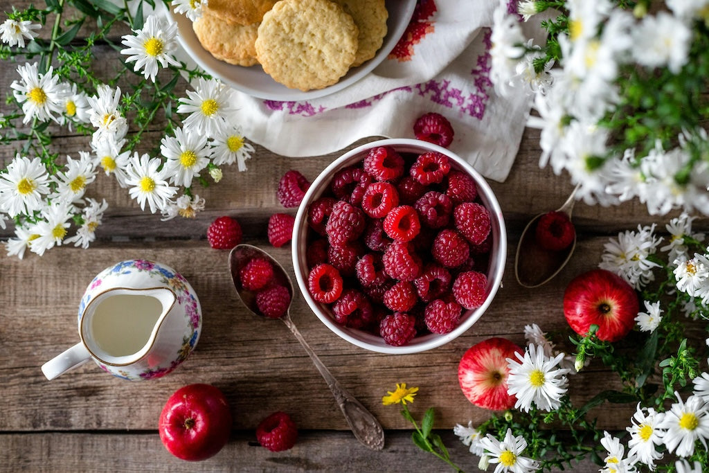 Auf einem Holztisch steht eine Schale mit frischen Himbeeren, drumherum sind Äpfel, Kekse, ein Krug Sahne und weiße Blumen arrangiert.