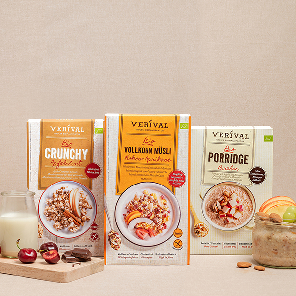 Drei Müslischachteln mit den Aufschriften Crunchy, Vollkorn Müsli und Porridge werden mit einem Glas, Kirschen und Mandeln auf beigem Hintergrund präsentiert.