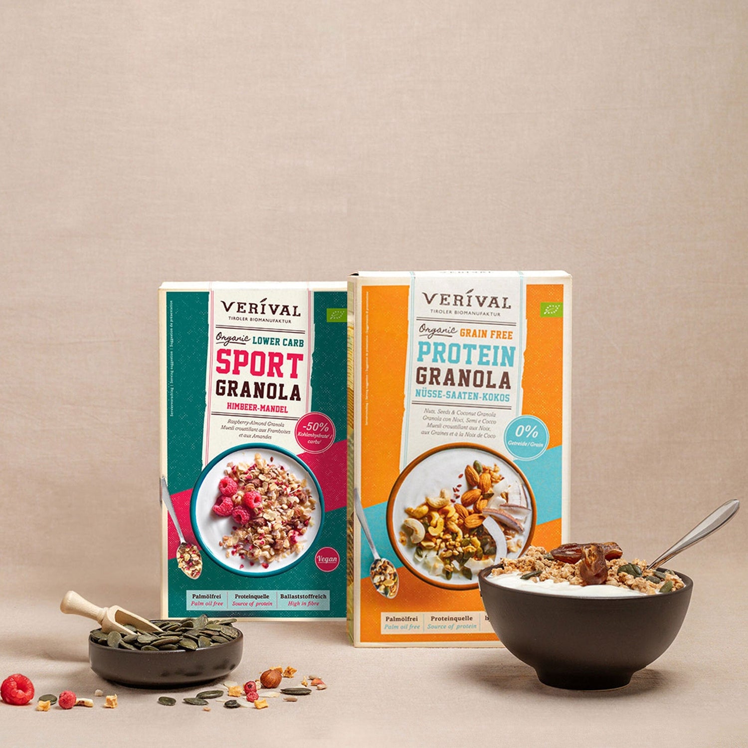 Zwei Schachteln Veríval-Granola, eine Sport-Granola und eine Protein-Granola, präsentiert mit einer Schale Granola, Körnern und Beeren auf beigem Hintergrund.