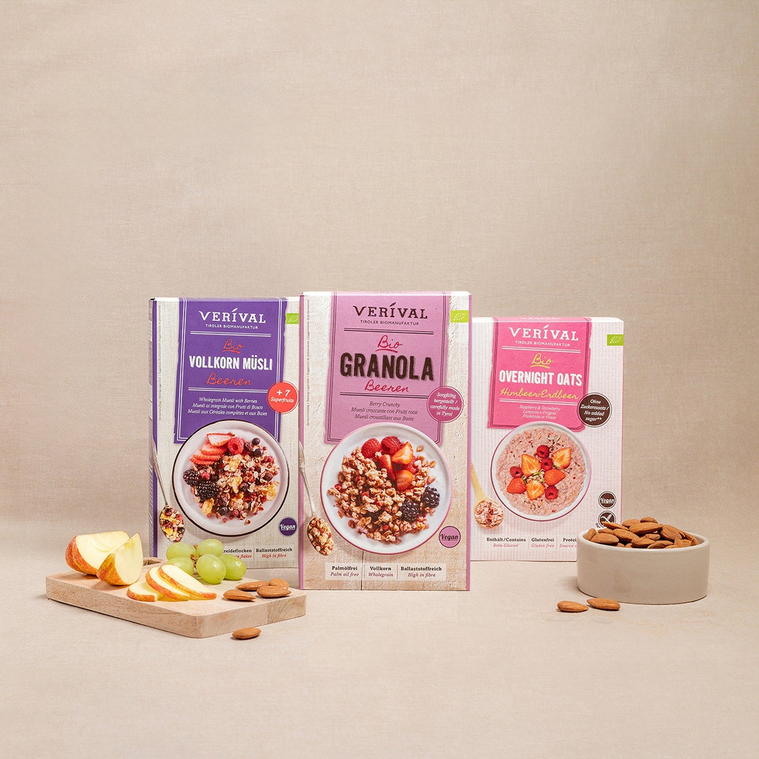 Drei Packungen Verival-Müsli (Vollkorn Müsli, Granola, Overnight Oats) werden mit einer Schale Mandeln und einem Schneidebrett mit Apfel- und Weintraubenscheiben präsentiert.