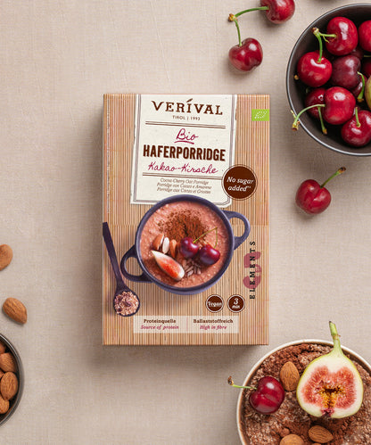 Eine Schachtel Verival Bio GmbH Bio Haferporridge Kirsche-Kakao steht auf einem hellen Tisch, umgeben von Kirschen, Feigen, Mandeln und einer Schüssel mit ballaststoffreichem Porridge - perfekt für ein warmes Frühstück.