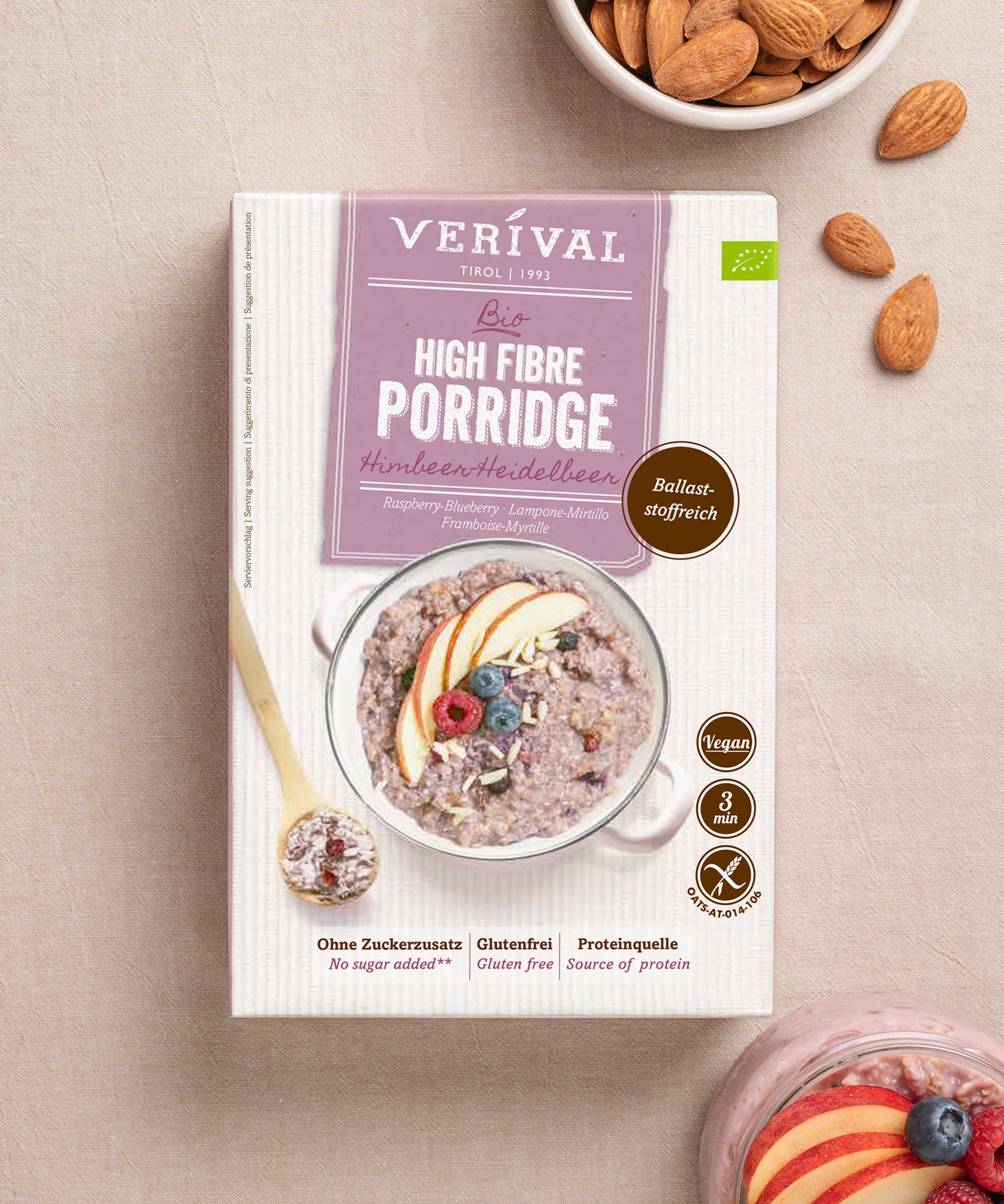 Der Bio Prebiotic Porridge Himbeer-Heidelbeer der Verival Bio GmbH auf einer leichten Unterlage, umgeben von Mandeln und einer Schale ballaststoffreichem, veganem Porridge, garniert mit frischen Obstscheiben - das ideale gesunde Frühstück.