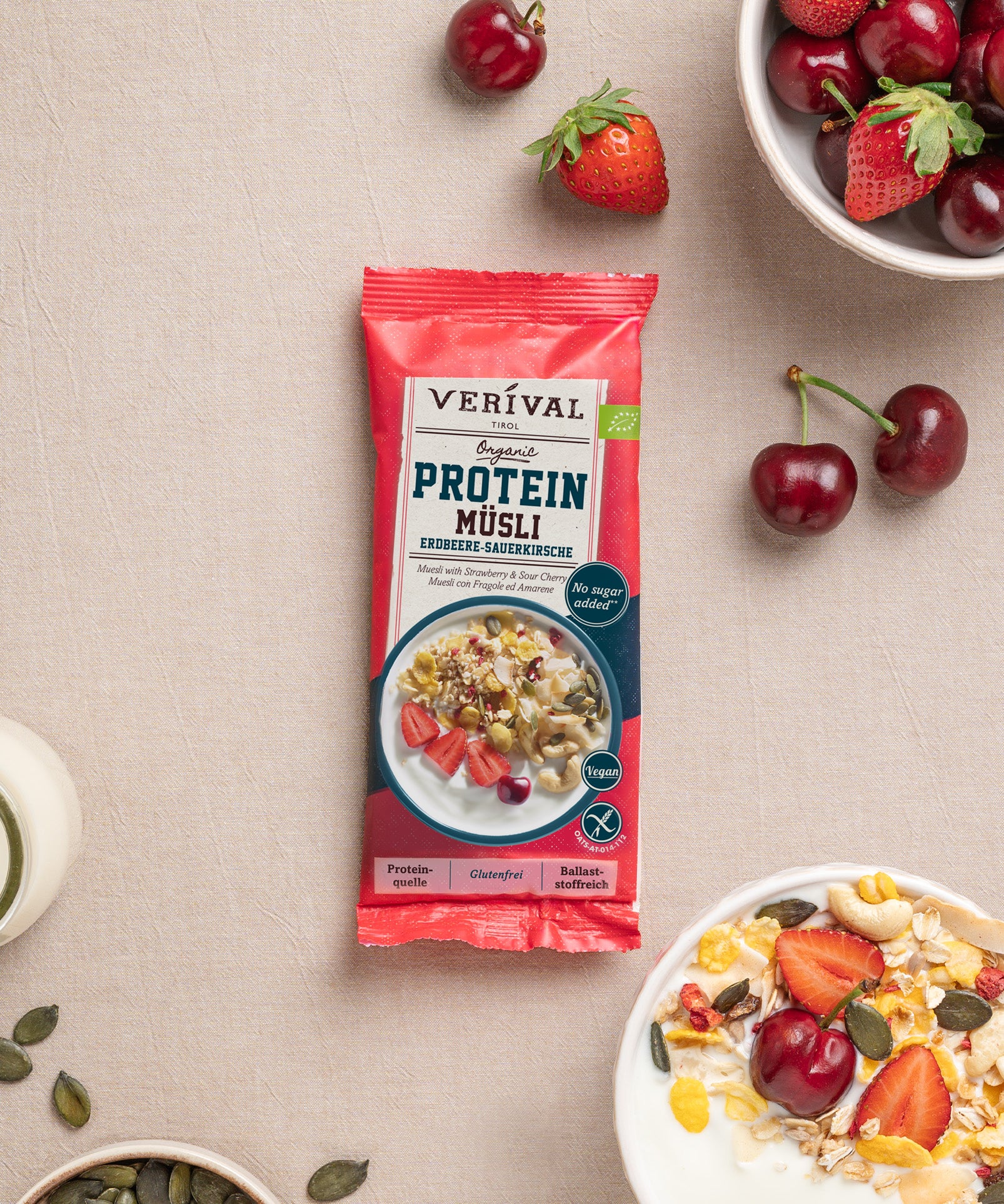 Eine Packung Protein Müsli Erdbeere-Sauerkirsche von Verival Bio liegt auf einer beigen Tischdecke, umgeben von einer Müslischale, frischen Erdbeeren, Kirschen, Kernen und Milch. 
