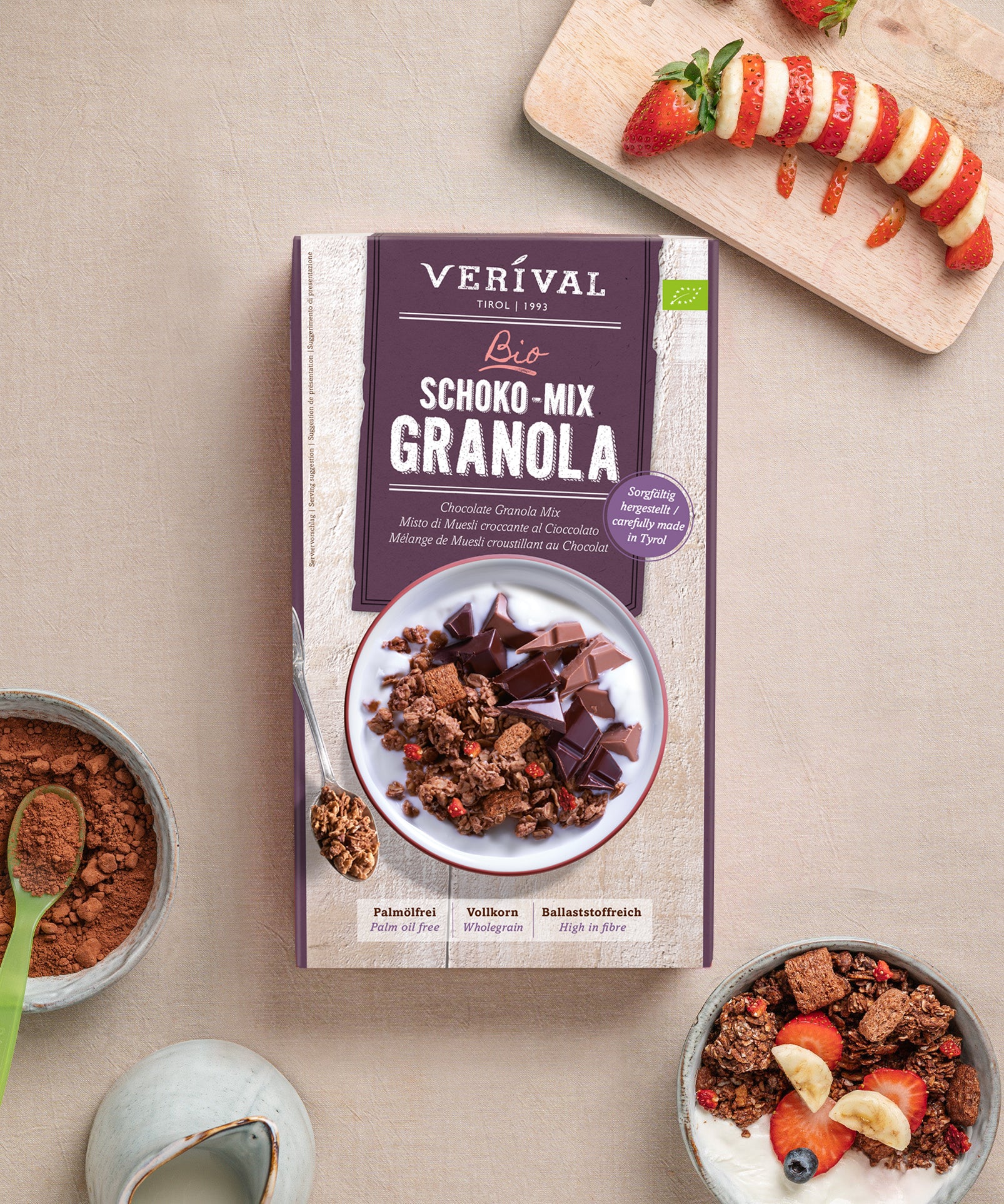 Eine Schachtel Verival Bio GmbH Bio Schoko Mix Granola steht auf einem Tisch neben einer Schüssel mit ballaststoffreichem Knusperfrühstück, daneben Obst, eine aufgeschnittene Erdbeere und ein Löffel.