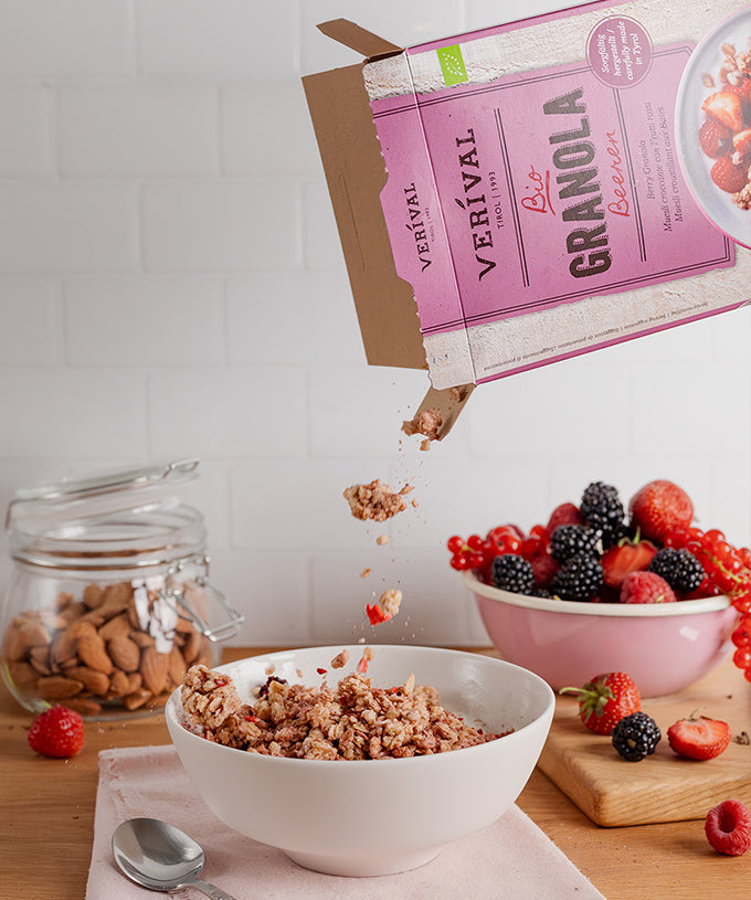 Eine Hand schüttet „Bio Beeren Granola“ der Verival Bio GmbH aus einer rosa Schachtel in eine weiße Schüssel. Mandeln, gemischte Beeren und weitere Beeren auf einem Schneidebrett umrahmen die Szene – ideal für ein knuspriges Frühstück. Ein Löffel liegt auf einer rosa Serviette.