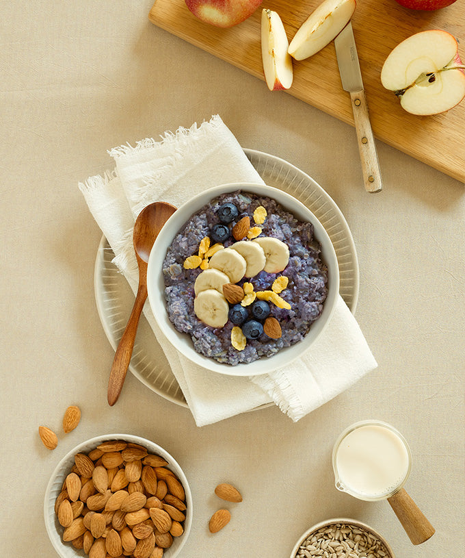 Eine Schüssel „Bio High Protein Porridge Heidelbeere-Banane“ von Verival Bio GmbH mit Banane, Heidelbeeren und Müsli wird mit einem Löffel serviert. Daneben liegen Mandeln, Apfelscheiben auf einem Brett, Sonnenblumenkerne und ein Krug Bio-Sojamilch.